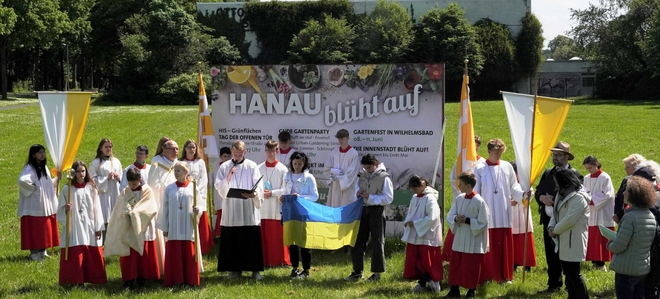 „Hanau blüht auf“ – Wie geschaffen war das Plakat am Wegrand auch als „himmlisches“  Thema für die dritte Station der Hanauer Prozession an Christi Himmelfahrt: Ukrainische Familien beteten hier  in ihrer Muttersprache mit mehreren hundert Gläubigen für den Frieden in ihrer Heimat. Mit dabei auch die ukrainische National-Flagge.